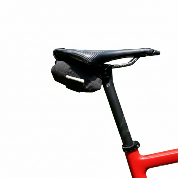 A black bicycle saddle bag mounted on a black saddle of a bicycle, photographed outdoors on a blurred green forest path.